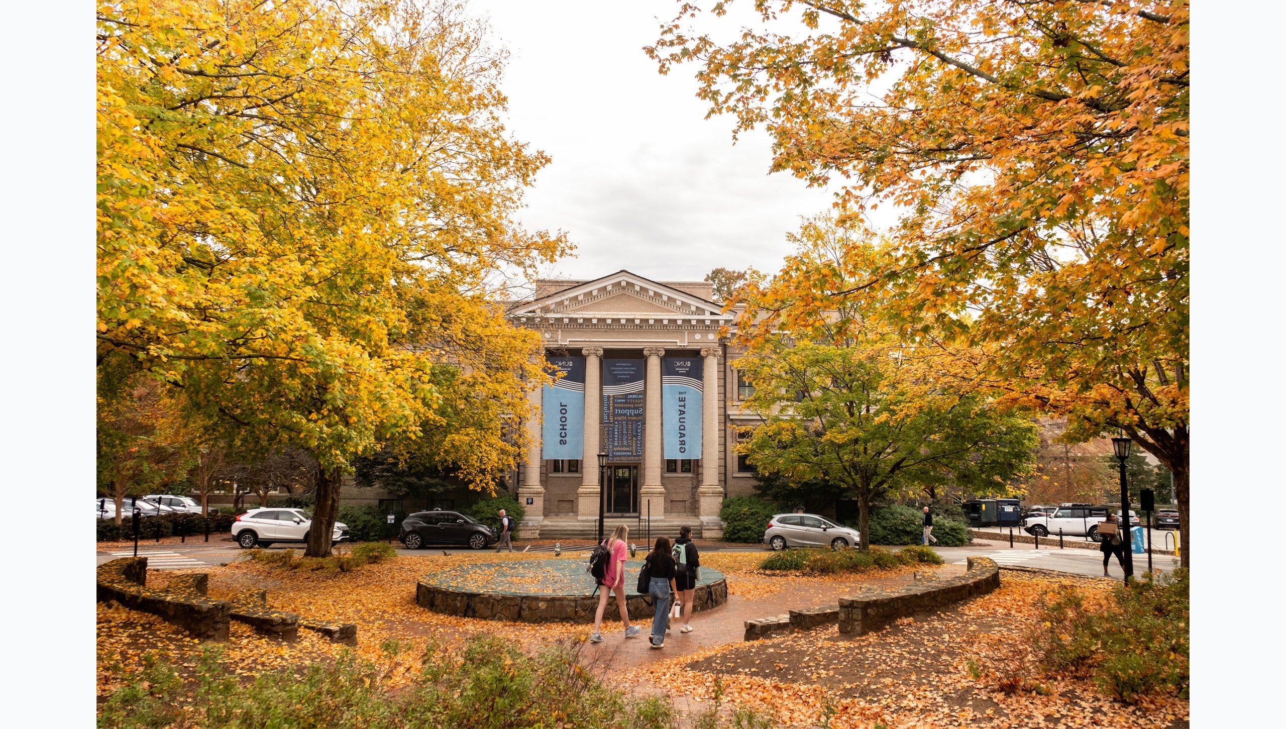 Students walking by the fountain in front of Bynum Hall on the campus of UNC-Chapel Hill. Trees with yellow and green leaves are seen in the background.